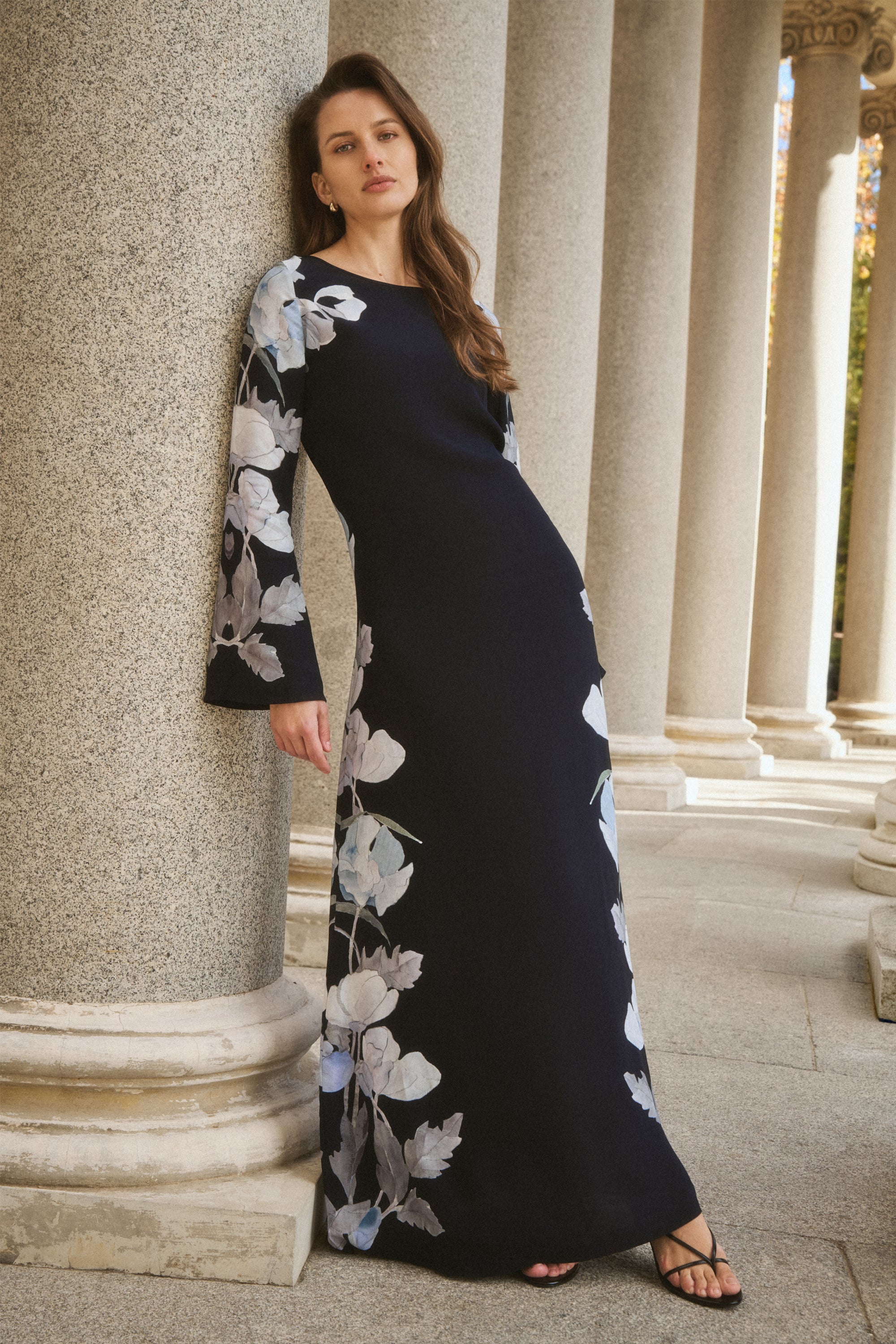 A woman wearing a long navy blue maxi dress with white and light blue floral prints on the sleeves and lower skirt, leaning against a large stone column in an outdoor colonnade.